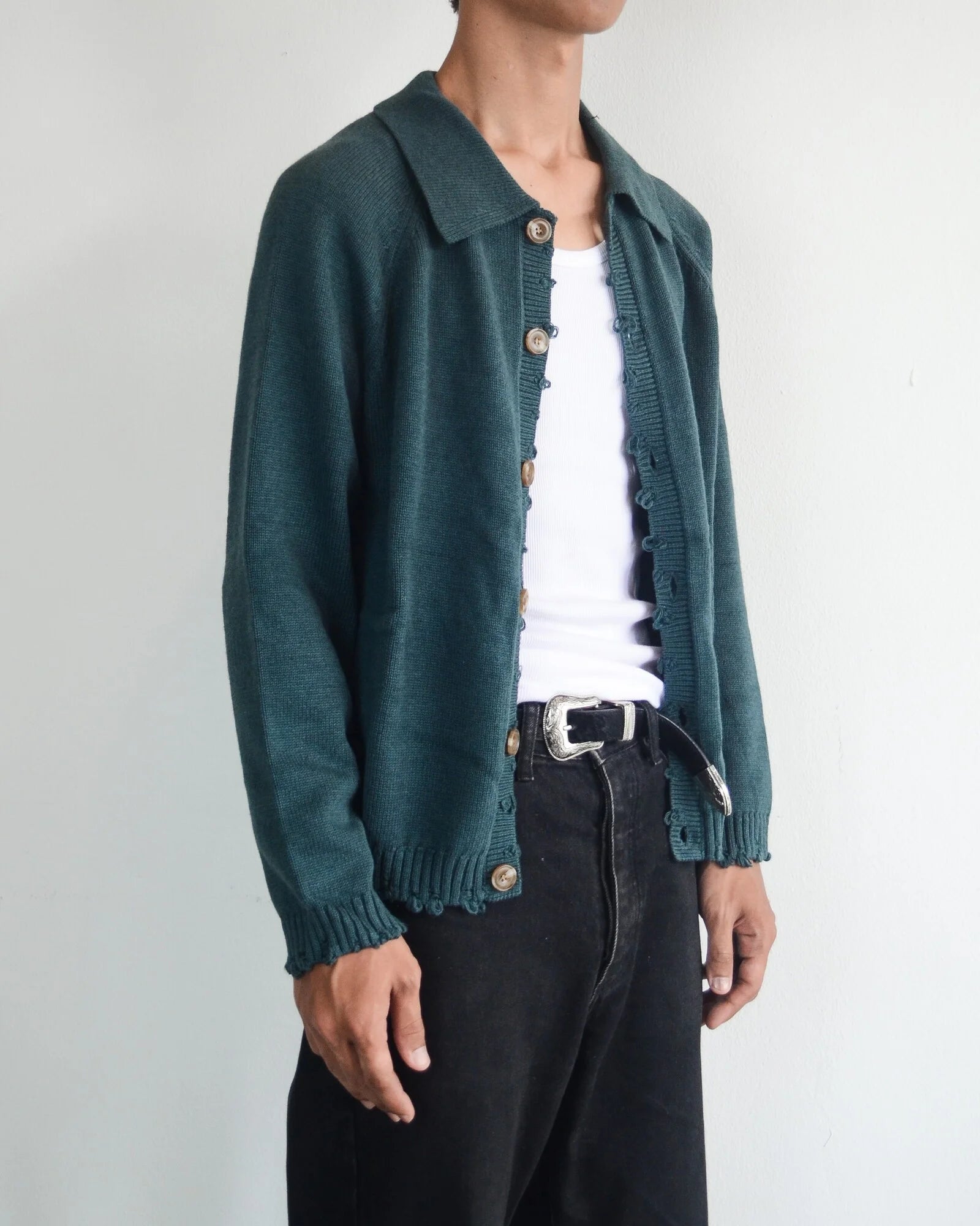 Person wearing a green cardigan over a white shirt with black pants on a plain background